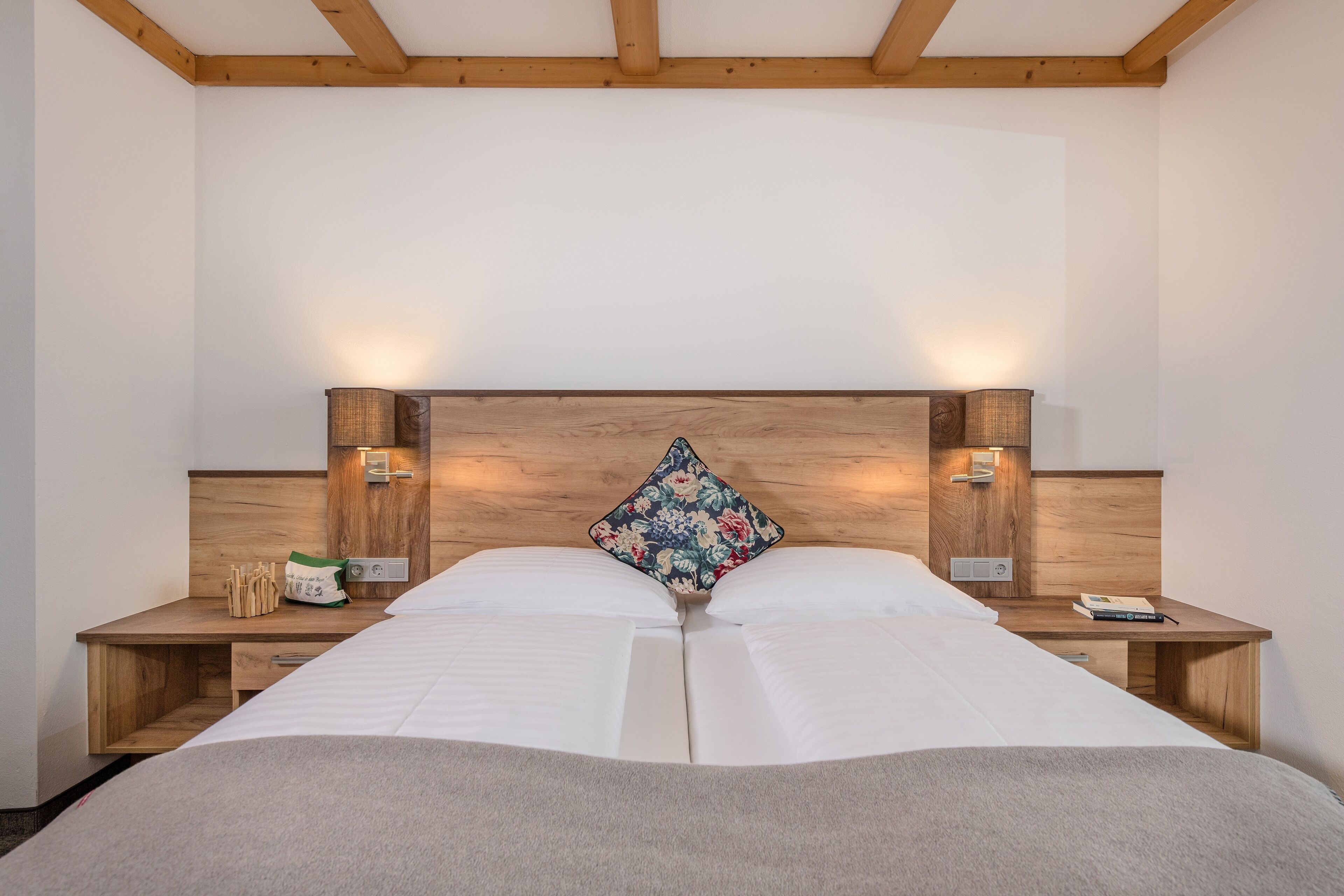 Una cama doble con cabecera de madera, dos mesitas de noche con lámparas y un cojín con estampado floral.