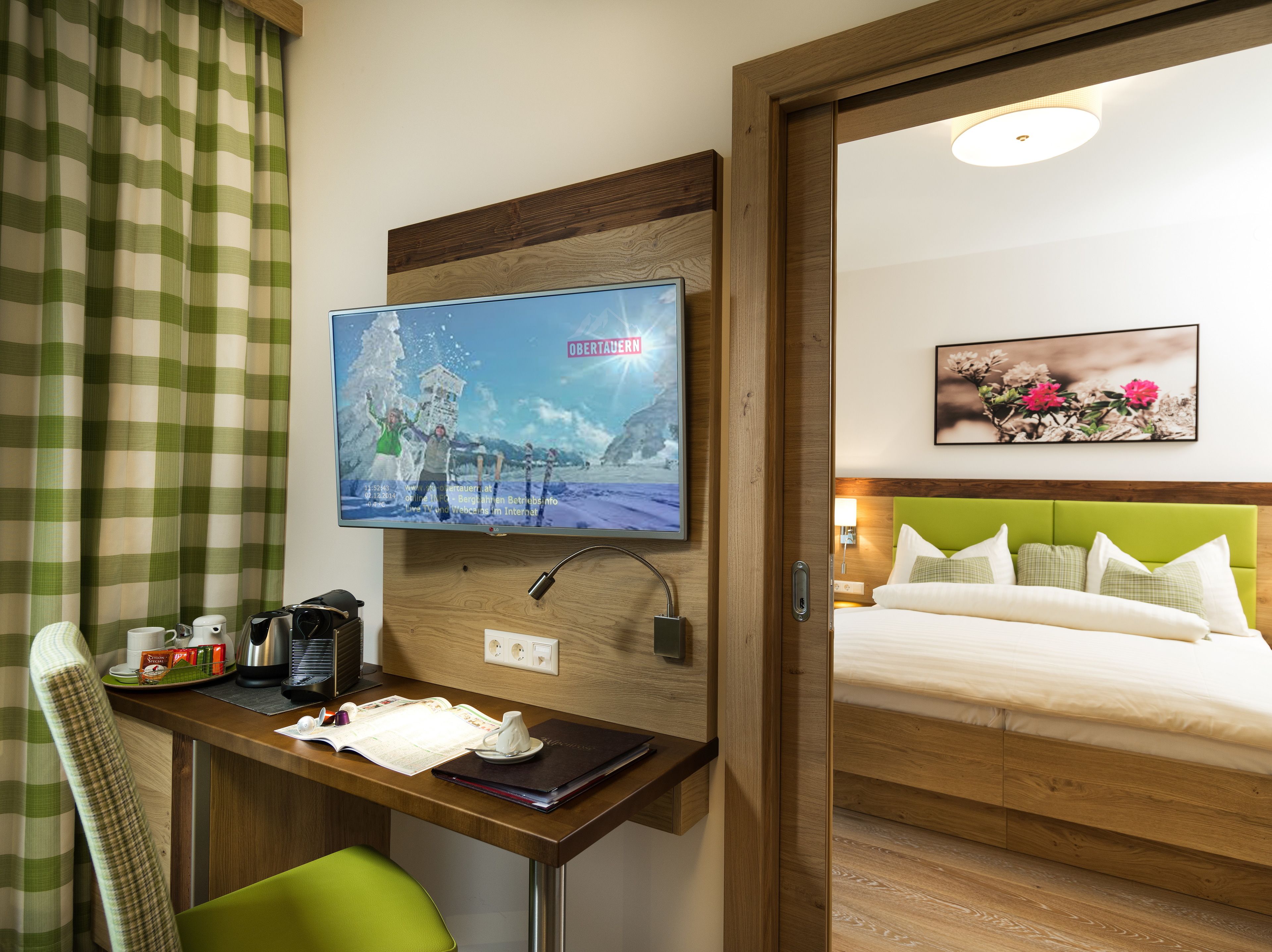 A hotel room with a wooden desk, a television displaying a snowy landscape, a bed with a green headboard, and a framed floral painting on the wall.