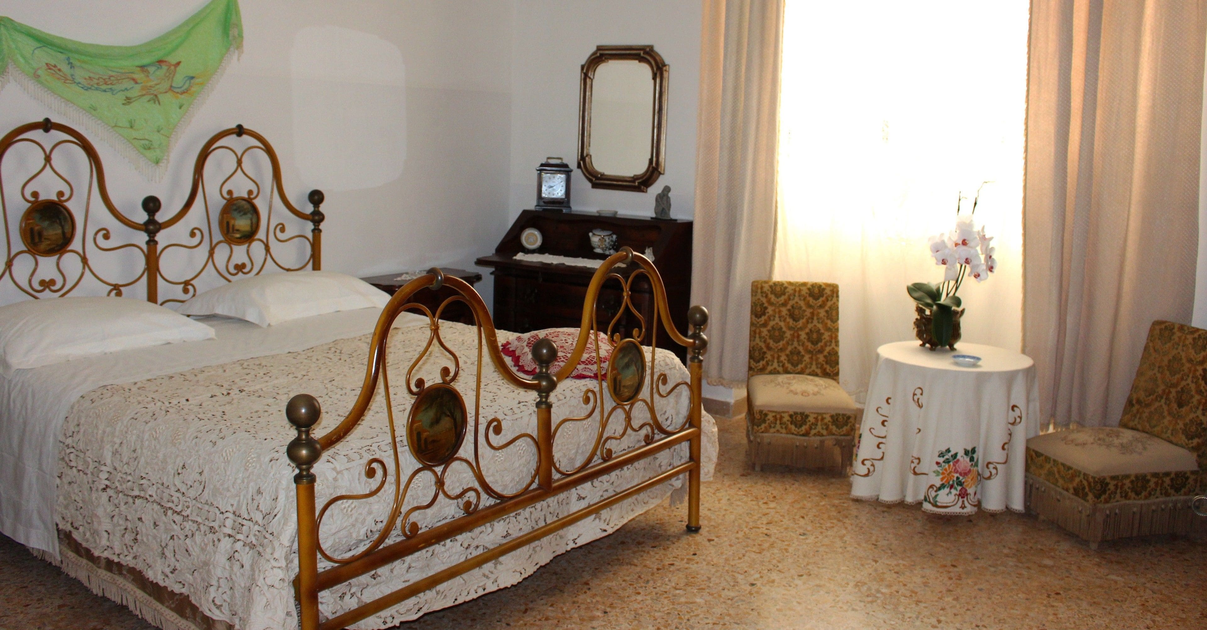 A bedroom with a four-poster bed, a small round table with a vase, two chairs, a nightstand with a clock, and a mirror on the wall.
