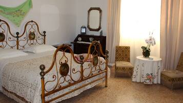 A bedroom with a four-poster bed, a small round table with a vase, two chairs, a nightstand with a clock, and a mirror on the wall.