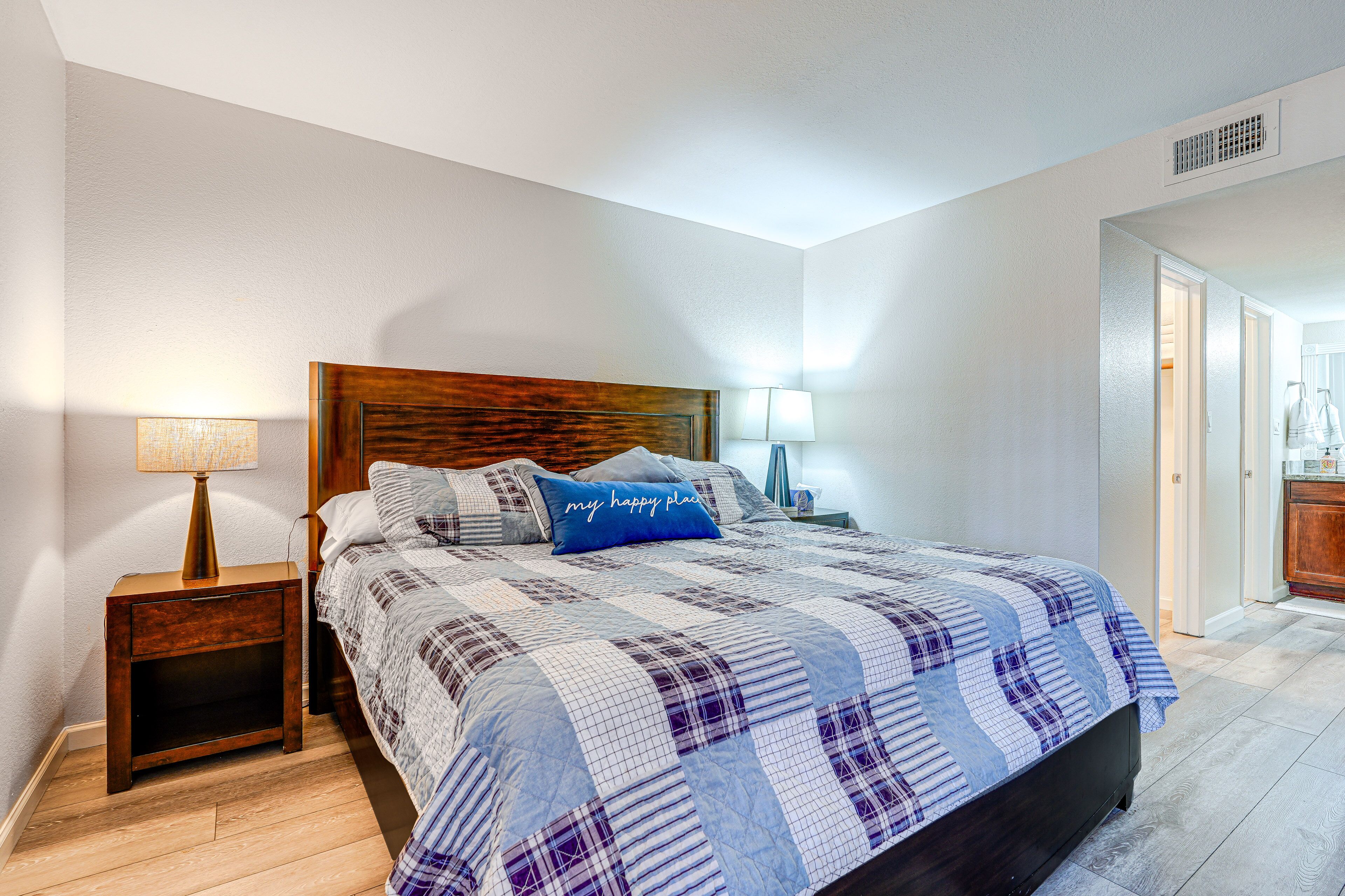 A bedroom with a wooden headboard, a plaid bedspread, a nightstand with a lamp, and a bedside table.