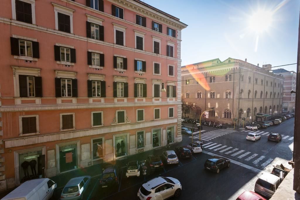 Un edificio a più piani con facciata rosa e persiane verdi, con vista sulla strada che mostra auto e un attraversamento pedonale.