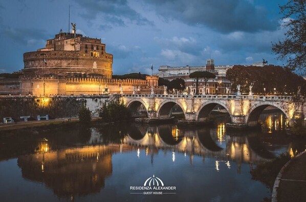 A historic building with a large central tower, a bridge with arches, and a body of water reflecting the structure. - Residenza Alexander (Rome)