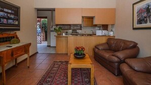 A living room with a brown leather sofa, a wooden coffee table with a flower vase, a television on a wooden stand, and a kitchen in the background. - Quail's Nest (Roodepoort)