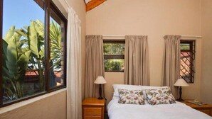A bedroom with a bed, two bedside tables, a window with curtains, and a view of greenery outside. - Quail's Nest (Roodepoort)