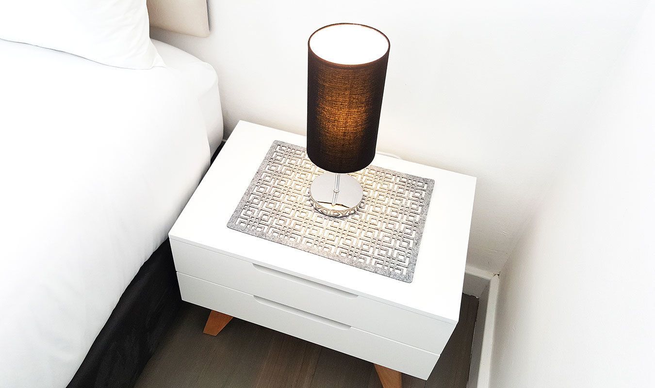 Uma mesa de cabeceira branca com uma luminária, um tapete estampado e uma gaveta.