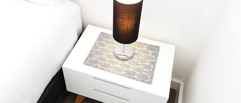 Uma mesa de cabeceira branca com uma luminária, um tapete estampado e uma gaveta.