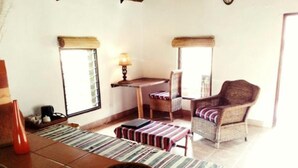 A cozy room with a tiled floor, a striped sofa, a wicker chair, a small table with a lamp, and a window with blinds. - Barefoot Lodge and Safaris - Malawi - Campsite (Chitedze)