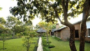 A garden with a paved pathway leading to a building with a brick facade and a thatched-roof structure. - Barefoot Lodge and Safaris - Malawi - Campsite (Chitedze)