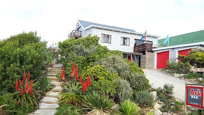 A two-story house with a red garage, a balcony, and a garden with various plants and flowers. - The-Ark B & B (Pearly Beach)
