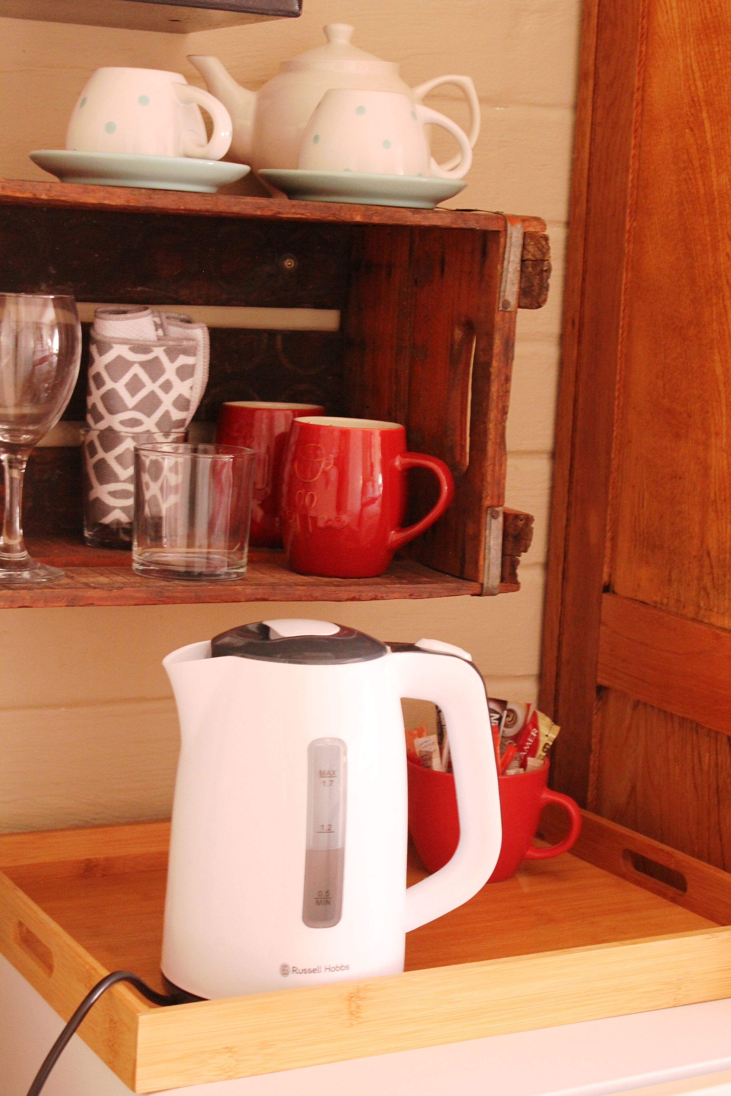 Un coin cuisine avec une bouilloire blanche posée sur un plan de travail en bois, une tasse rouge et une étagère contenant divers ustensiles de cuisine, notamment des théières et des tasses.