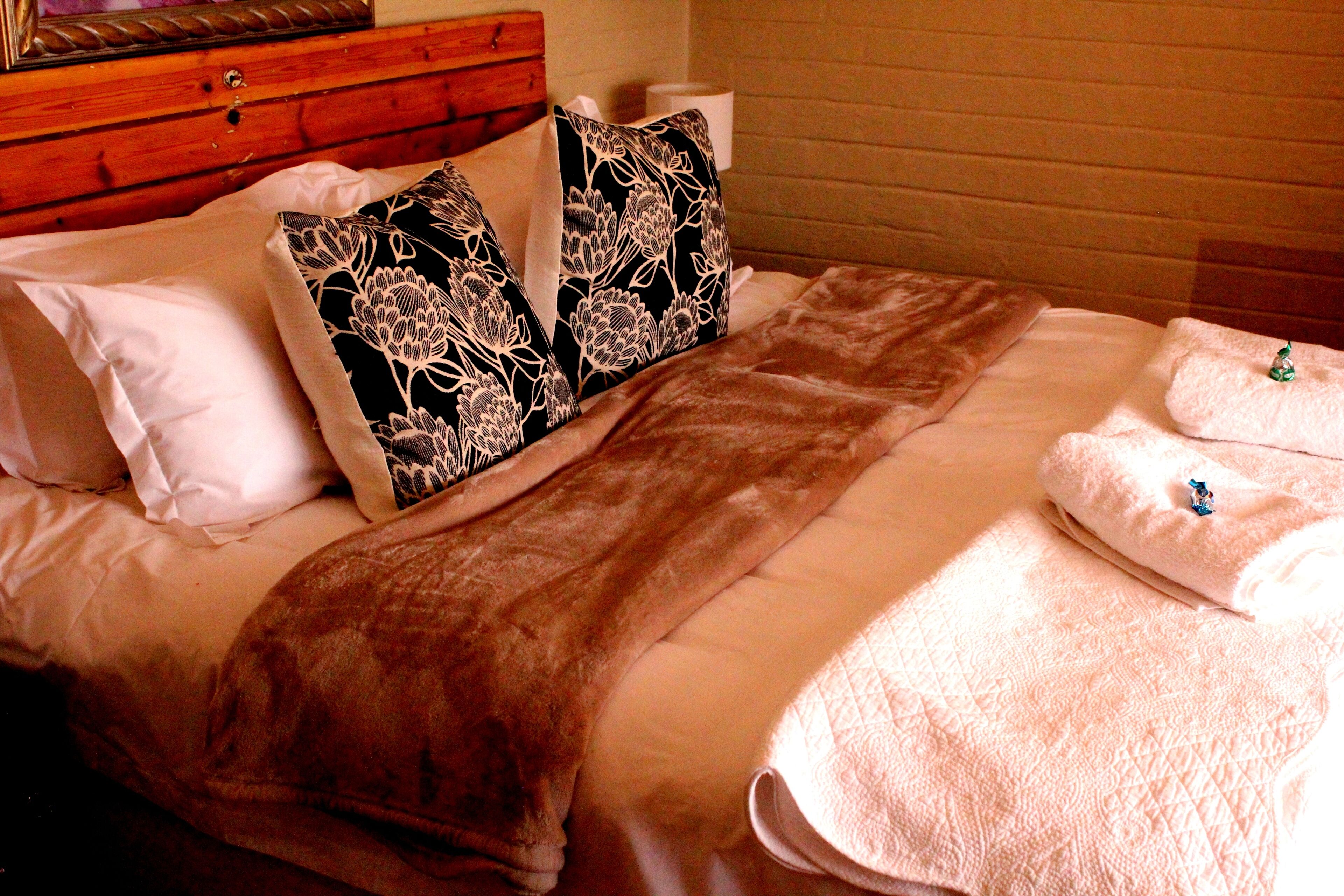 A neatly made bed with a brown blanket, two white pillows with black floral patterns, and a white towel on the side.