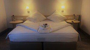 A neatly made bed with white linens, two bedside tables with lamps, and a small vase with flowers on one table. - A&M Hotel Barsinghausen (Barsinghausen)