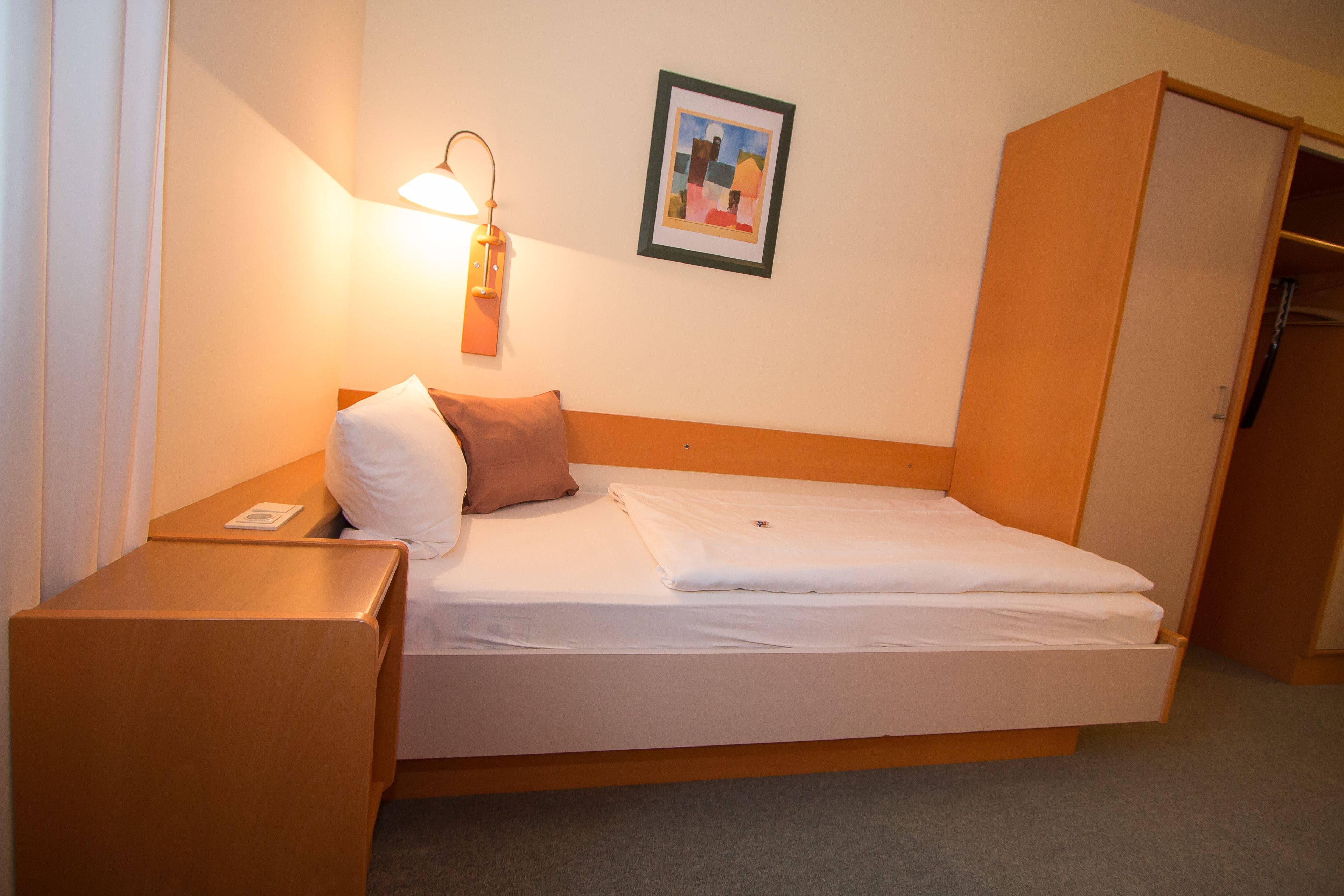 A neatly made bed with white linens, a brown pillow, and a bedside lamp.