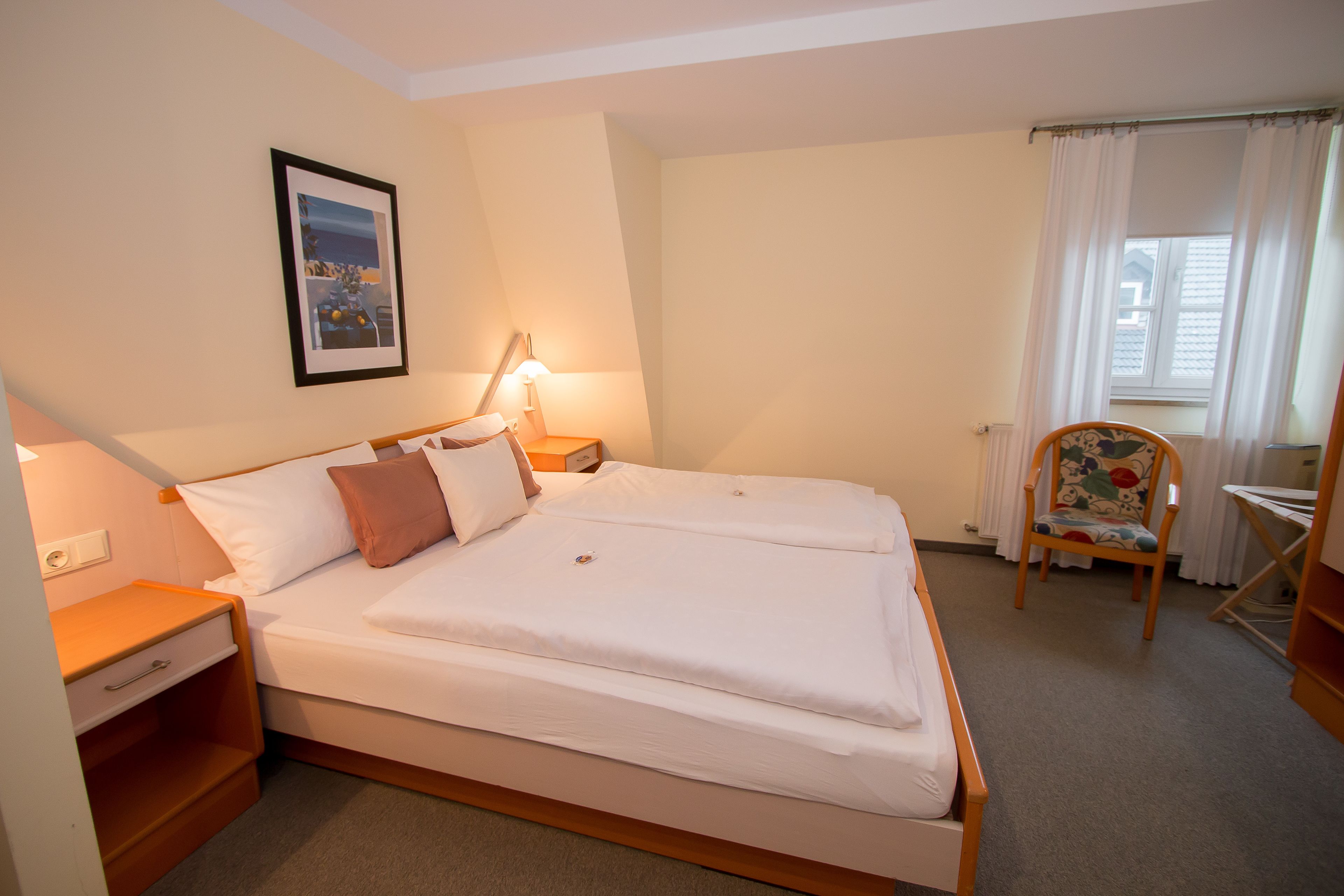 A neatly made bed with white linens, a bedside table with a lamp, a chair with a floral pattern, and a framed picture on the wall.