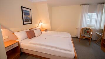 A neatly made bed with white linens, a bedside table with a lamp, a chair with a floral pattern, and a framed picture on the wall.