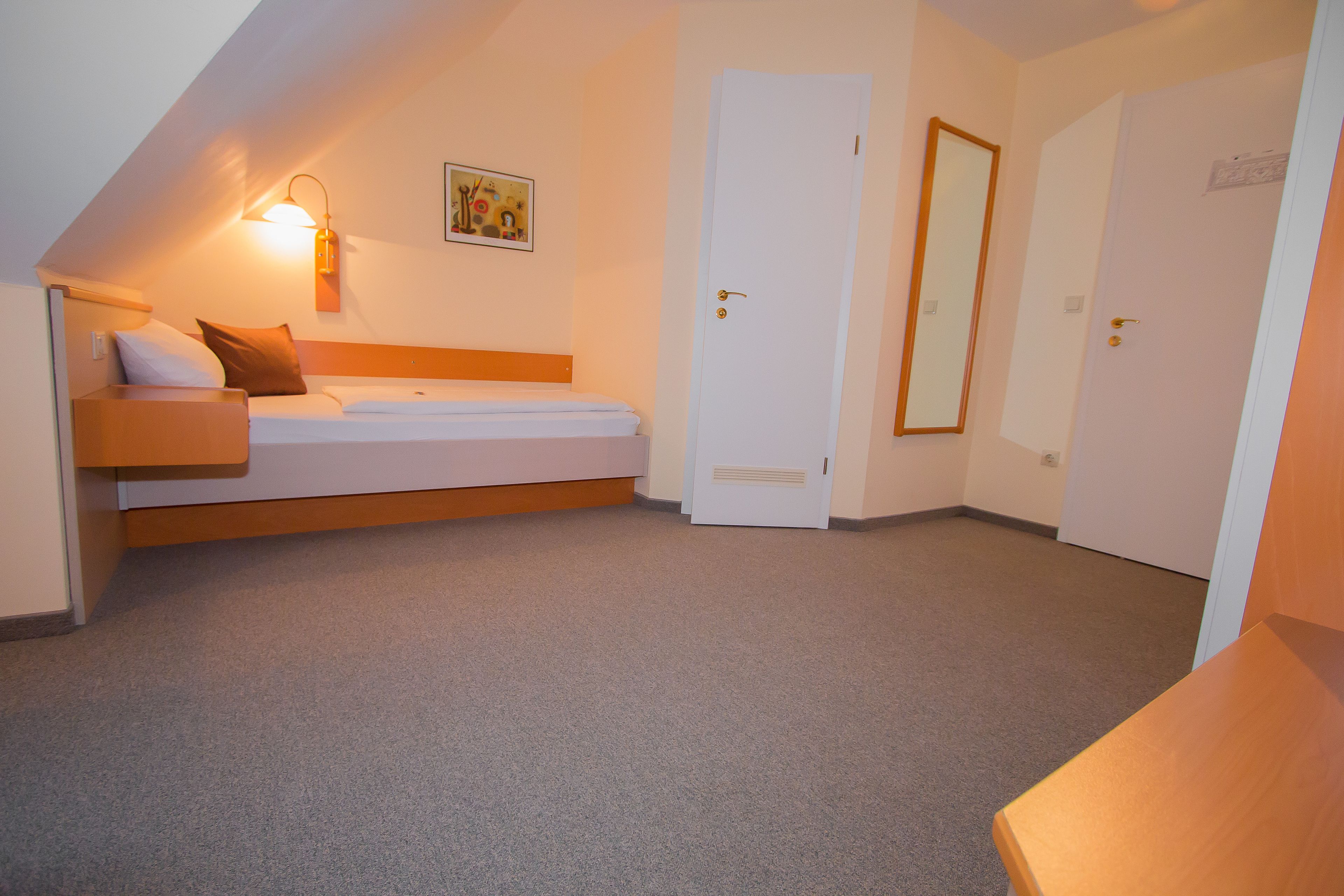 A small, well-lit room with a bed, a wall-mounted light, and a door.