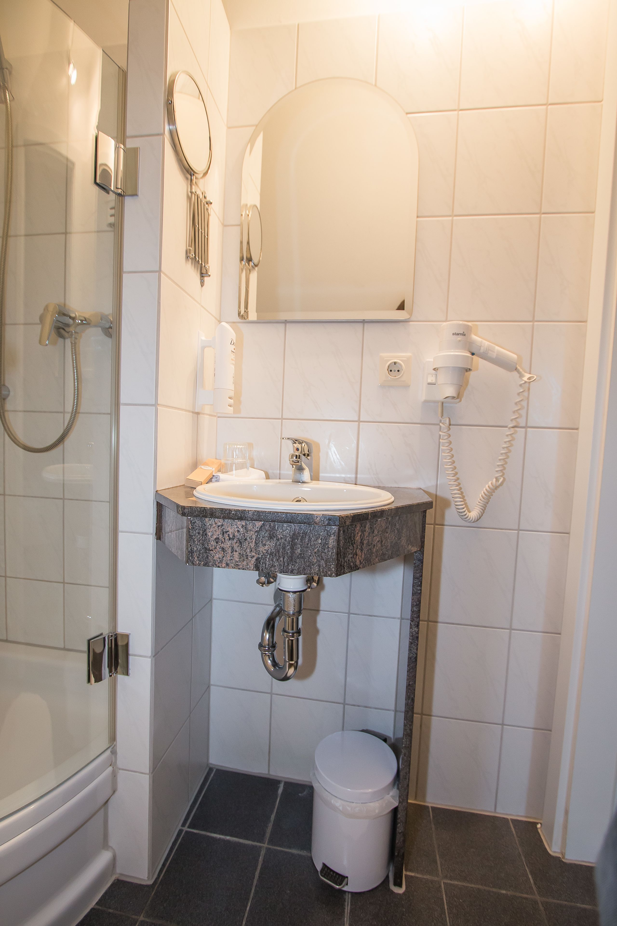 A bathroom with a glass-enclosed shower, a sink with a marble countertop, a toilet, and a hairdryer on the wall.