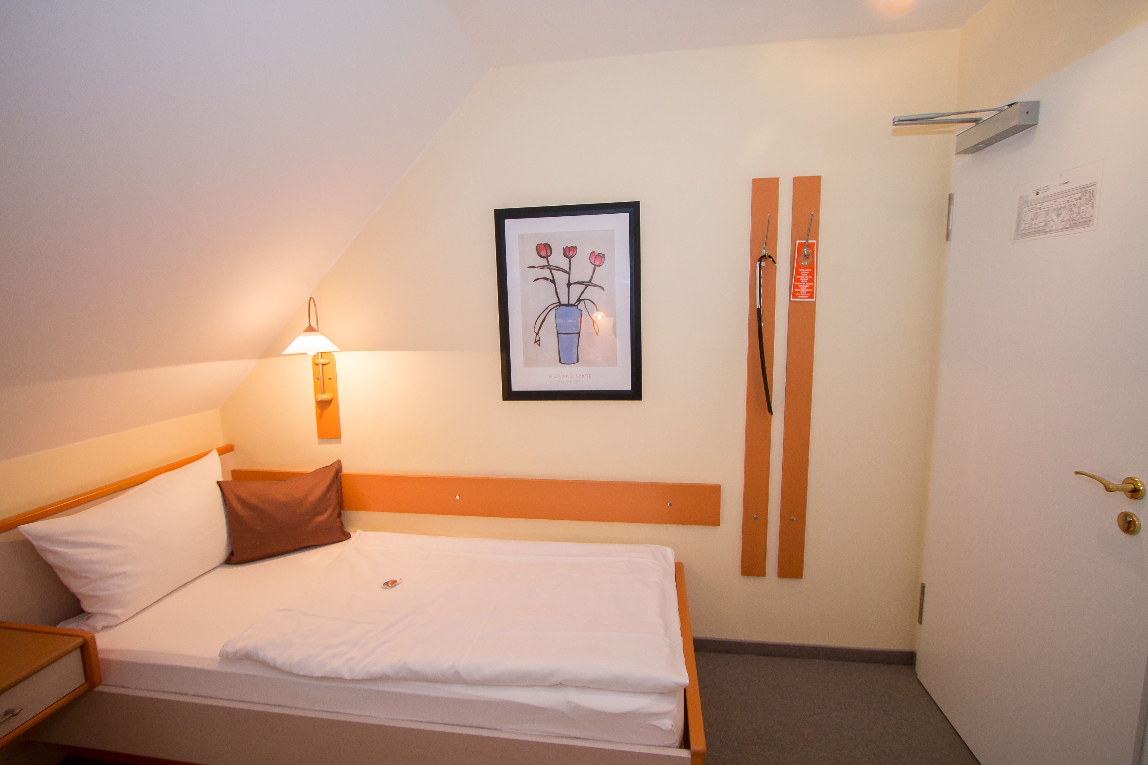 A hotel room with a bed, a nightstand, a wall-mounted lamp, a framed picture, and a door with a handle.
