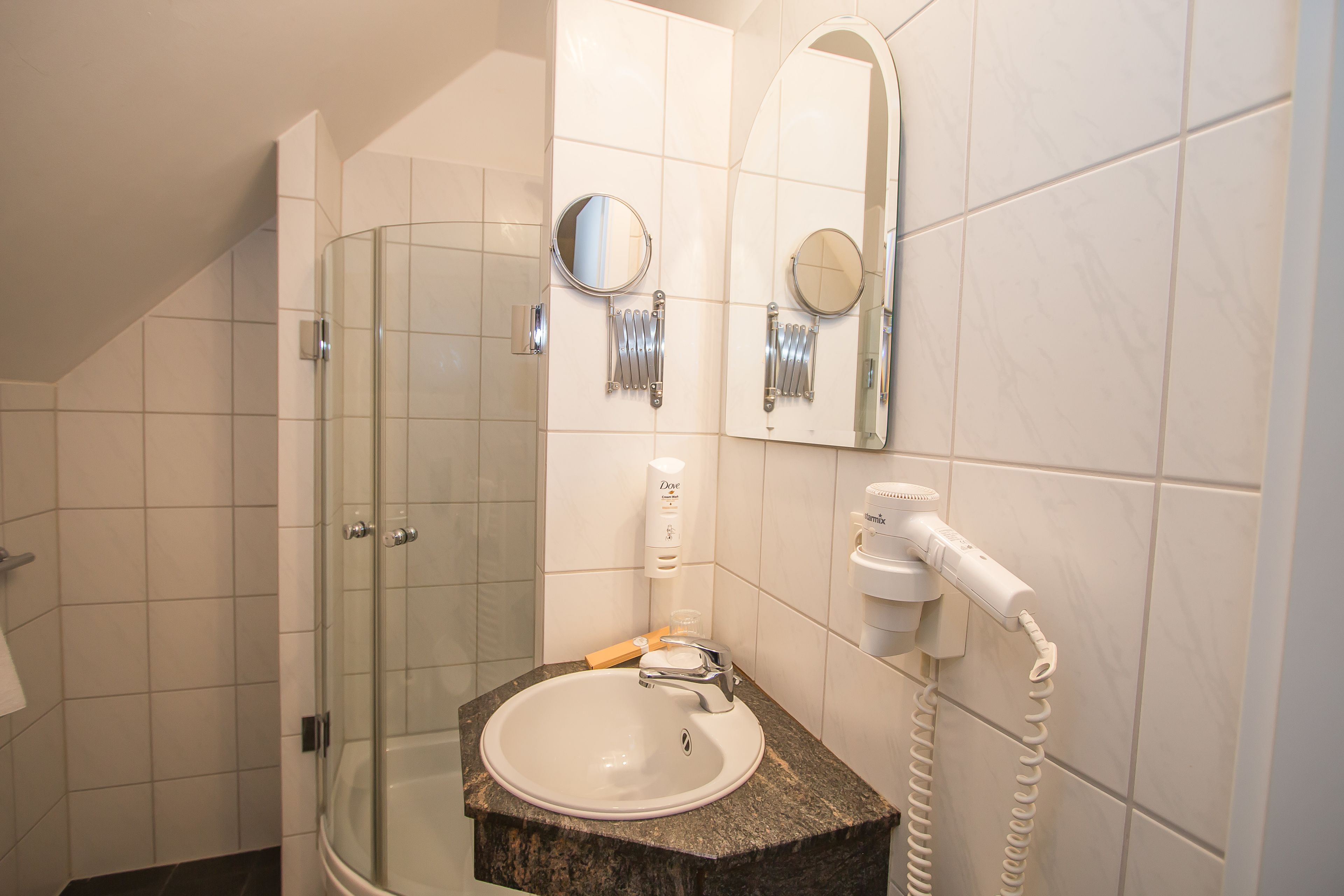 A bathroom with a glass shower enclosure, a sink with a marble countertop, two wall-mounted mirrors, and a hairdryer on the wall.