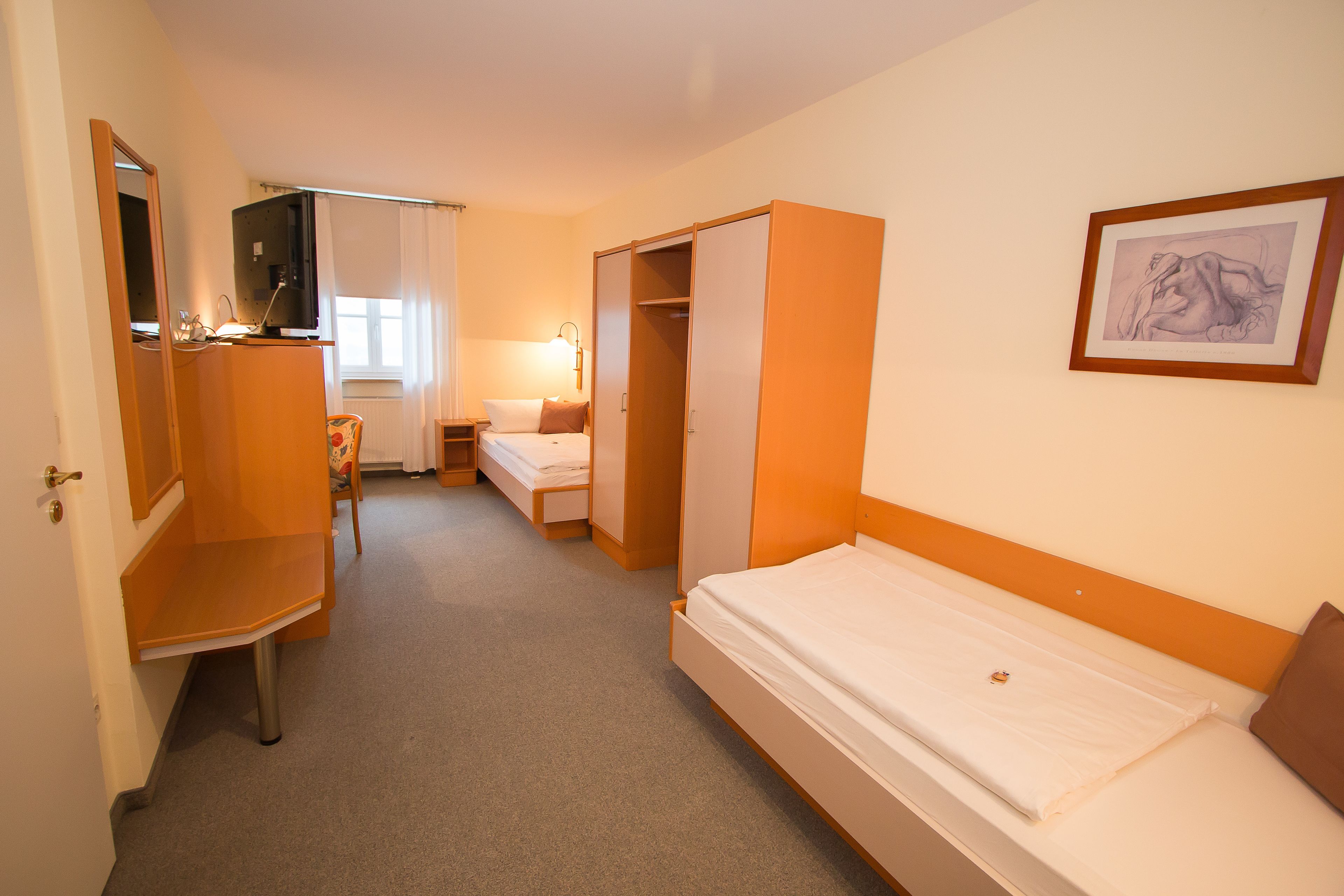 A hotel room with a bed, a desk, a chair, a wardrobe, and a framed picture on the wall.