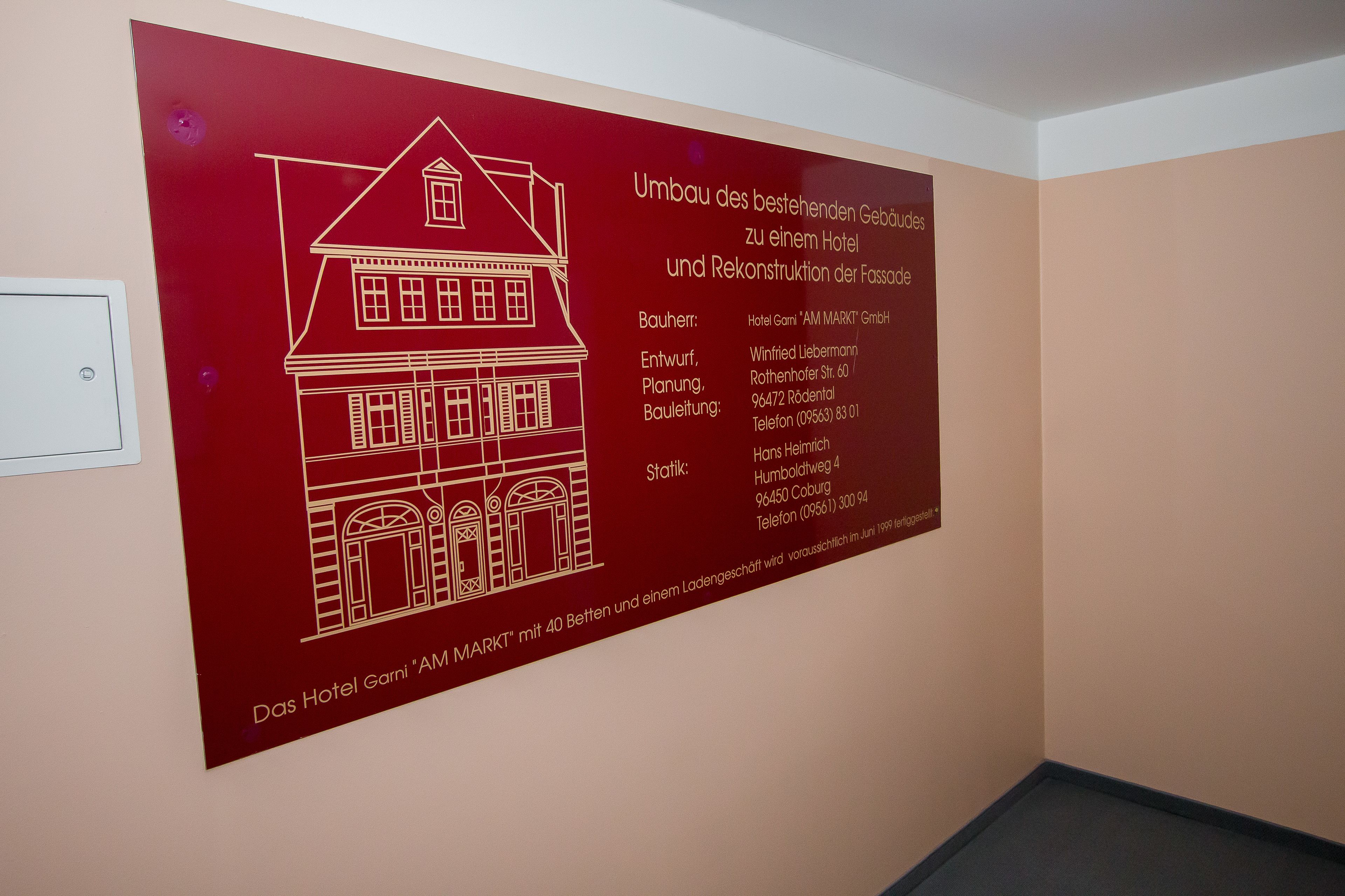 Eine Wand mit einem roten Poster, das die Kontur eines Gebäudes zeigt und Text über Bau und Restaurierung eines Hotels sowie der Fassade enthält.
