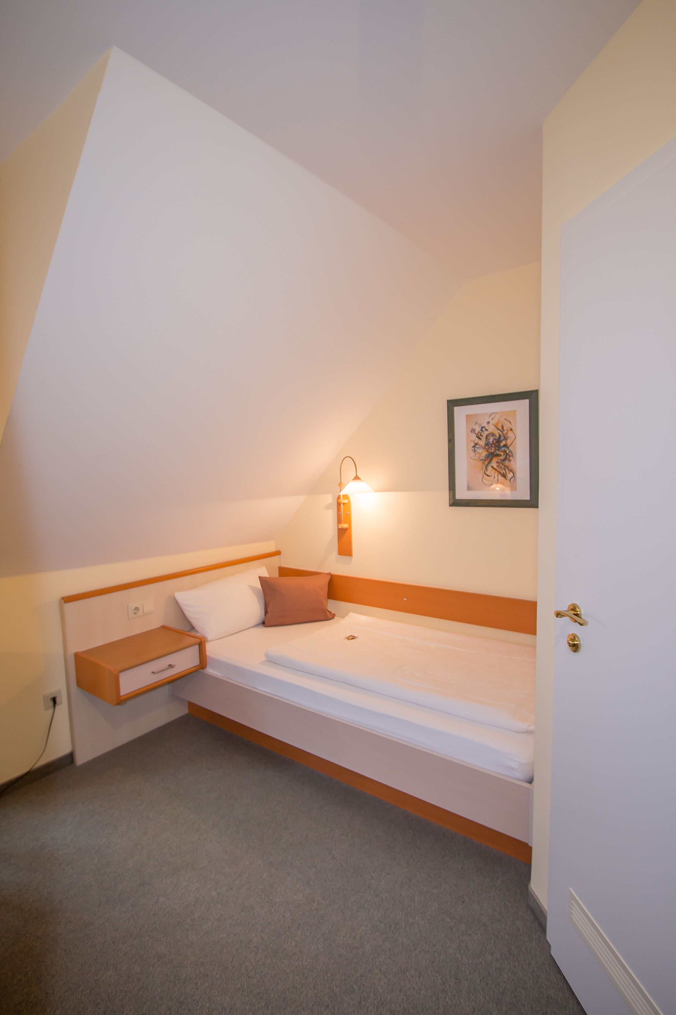 A single bed with a wooden headboard, a bedside table with a drawer, a wall-mounted light, and a framed picture on the wall.