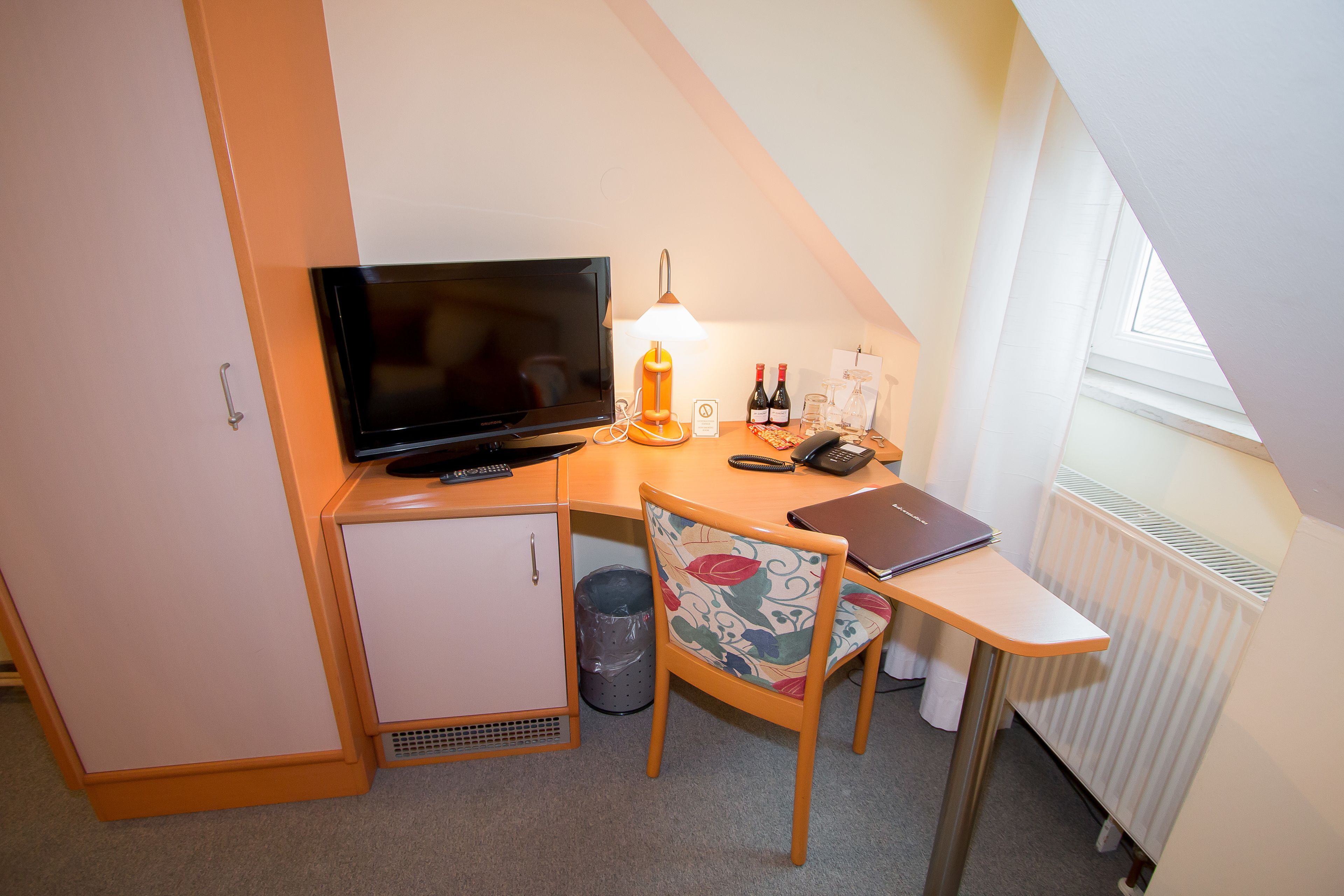 Une pièce compacte et bien éclairée, comprenant un bureau, une chaise, un téléviseur, une lampe et un petit meuble.