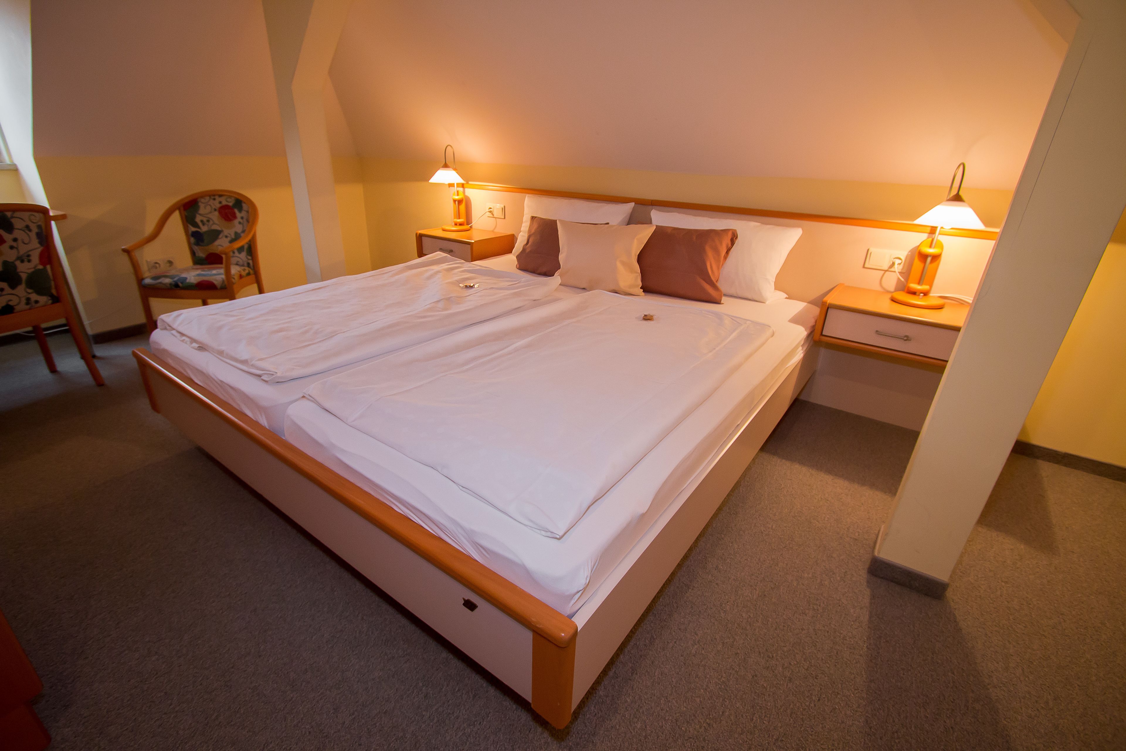A double bed with a wooden frame, two bedside tables with lamps, a chair with a floral pattern, and a carpeted floor.