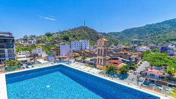 A swimming pool with a tiled deck, surrounded by buildings and a clock tower.