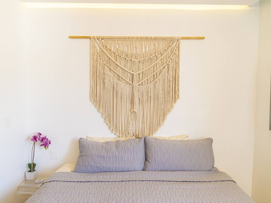 Un lit recouvert d’une couvre-lit à motifs géométriques, une décoration murale en macramé au-dessus, et une petite orchidée en pot posée sur une table de chevet.