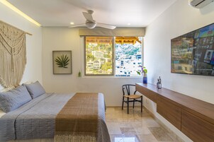Un dormitorio con cama, escritorio y ventana con vista a edificios.