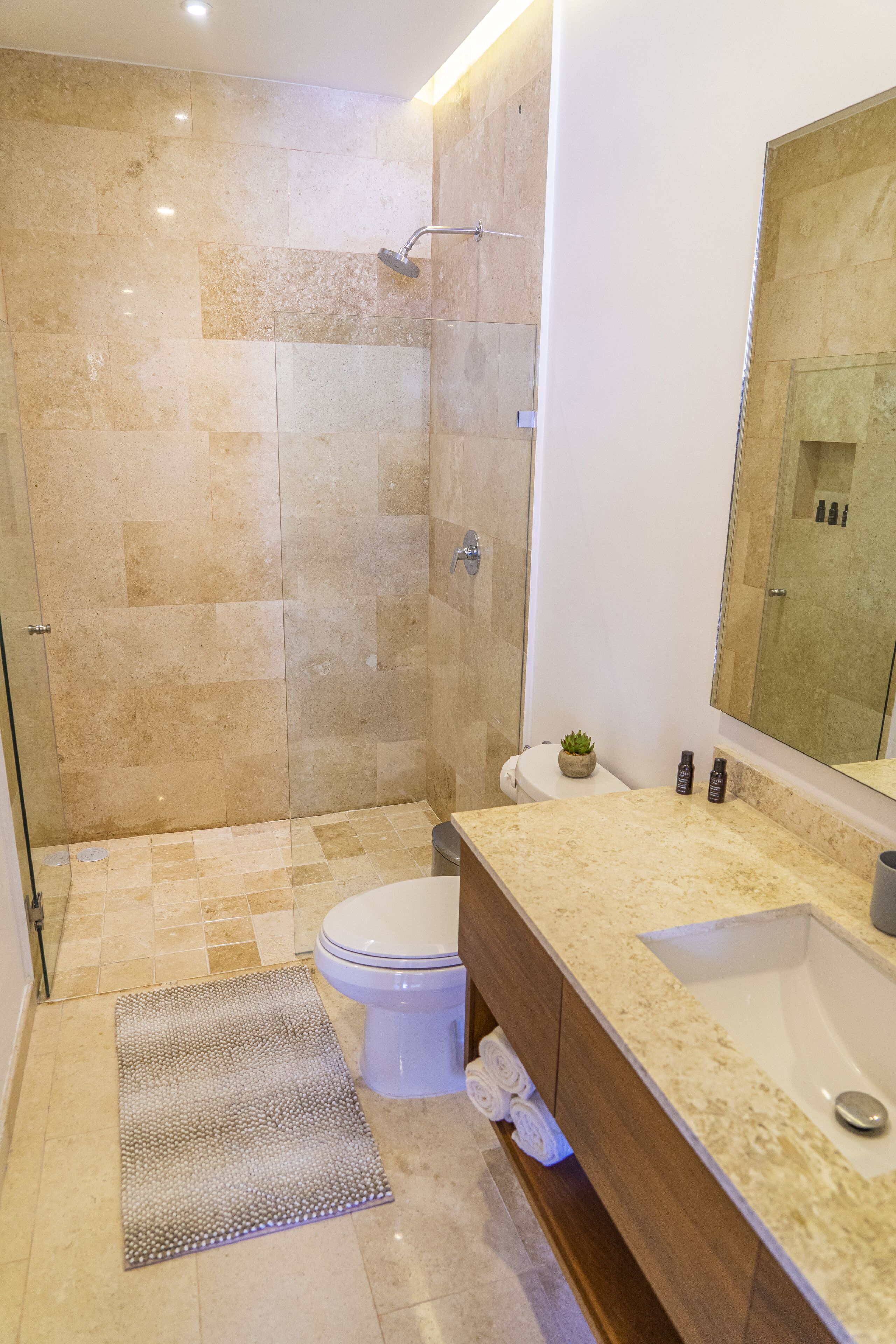 Un baño moderno con ducha de vidrio, inodoro blanco, encimera beige con lavamanos y espejo sobre el lavamanos.