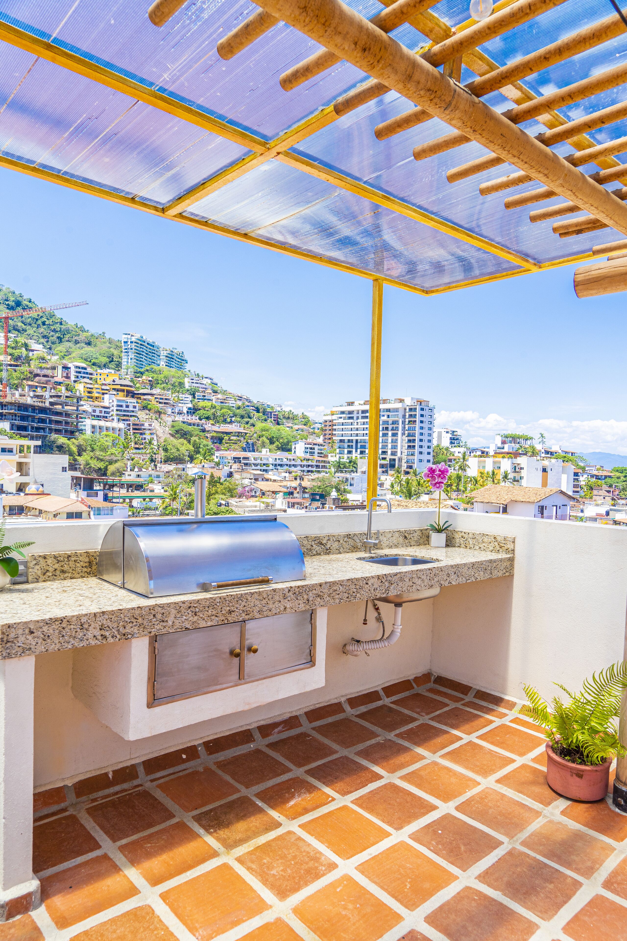 Una zona de cocina exterior con una parrilla, pileta y plantas en macetas en una terraza con vista a la ciudad.