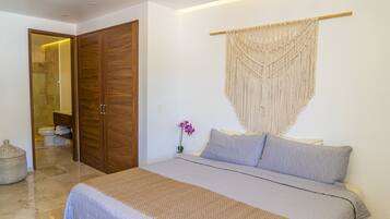 Un dormitorio con cama, armario de madera, un colgador de macramé y un jarrón pequeño con una flor.