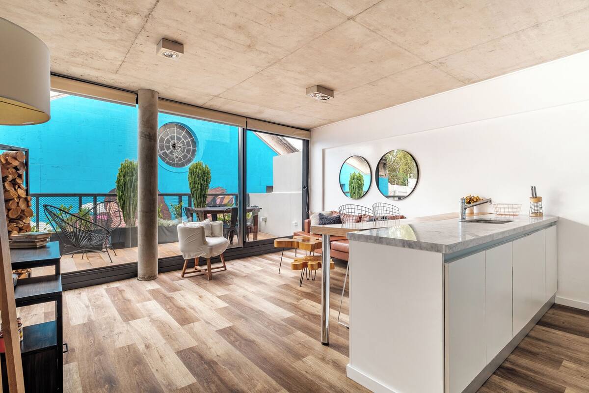 A modern kitchen with a large island, wooden flooring, and large windows offering a view of the outdoors.