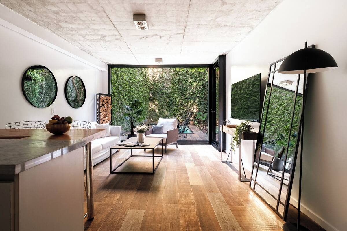 A modern living room with a wooden floor, a sofa, a coffee table, and a dining area with a bowl of fruit. There are two round mirrors on the wall and a large window with a view of greenery.