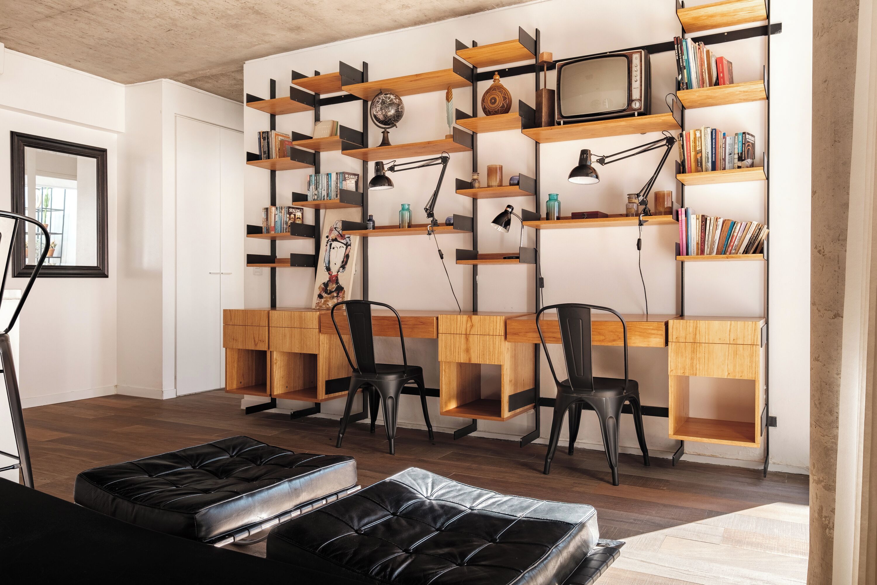 A modern workspace with wooden desks, black chairs, and a black leather ottoman. Shelves with books and decorative items are mounted on the wall.
