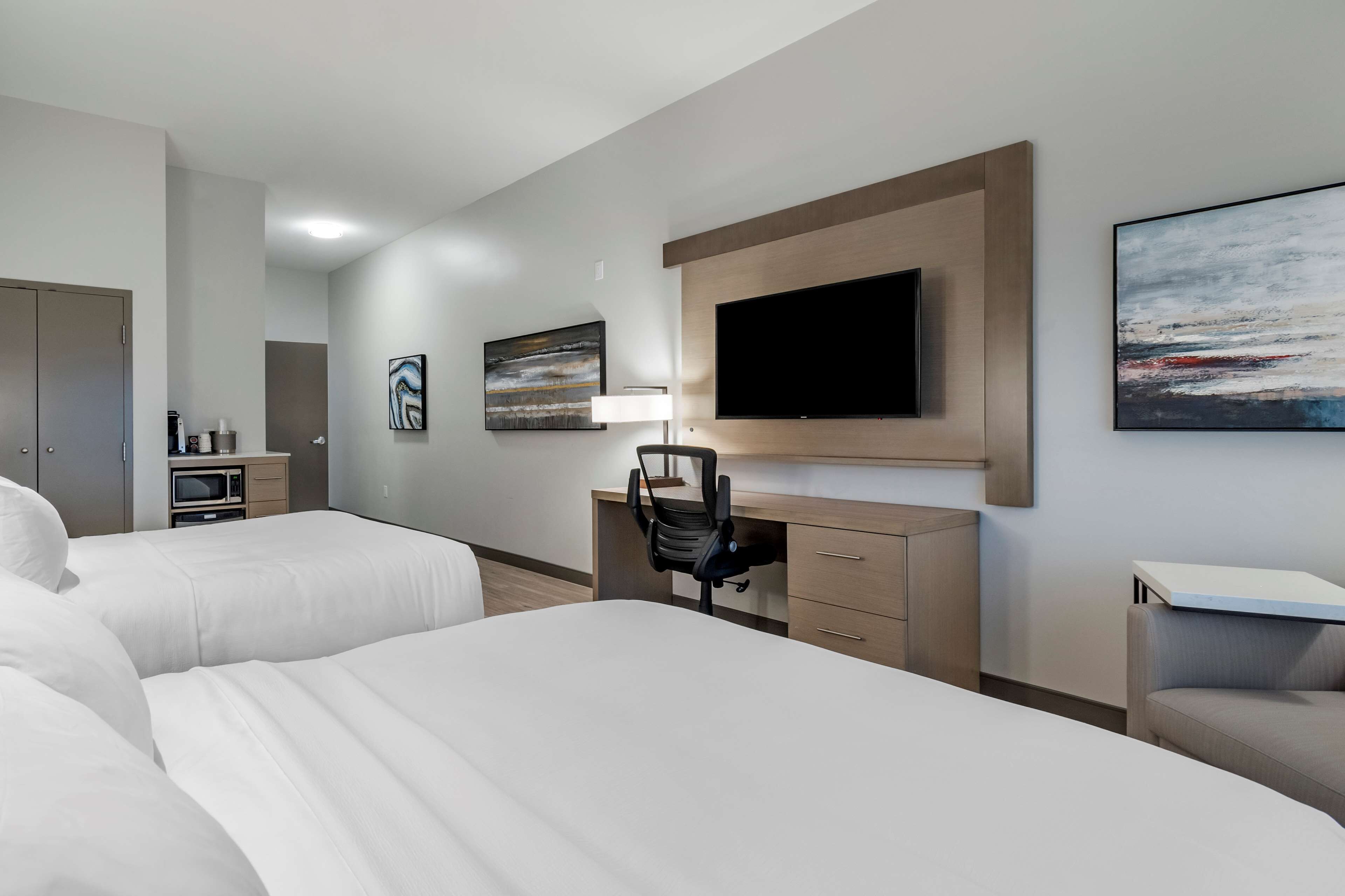 Habitación de hotel con dos camas, un escritorio, una silla, un televisor y obras de arte en las paredes.