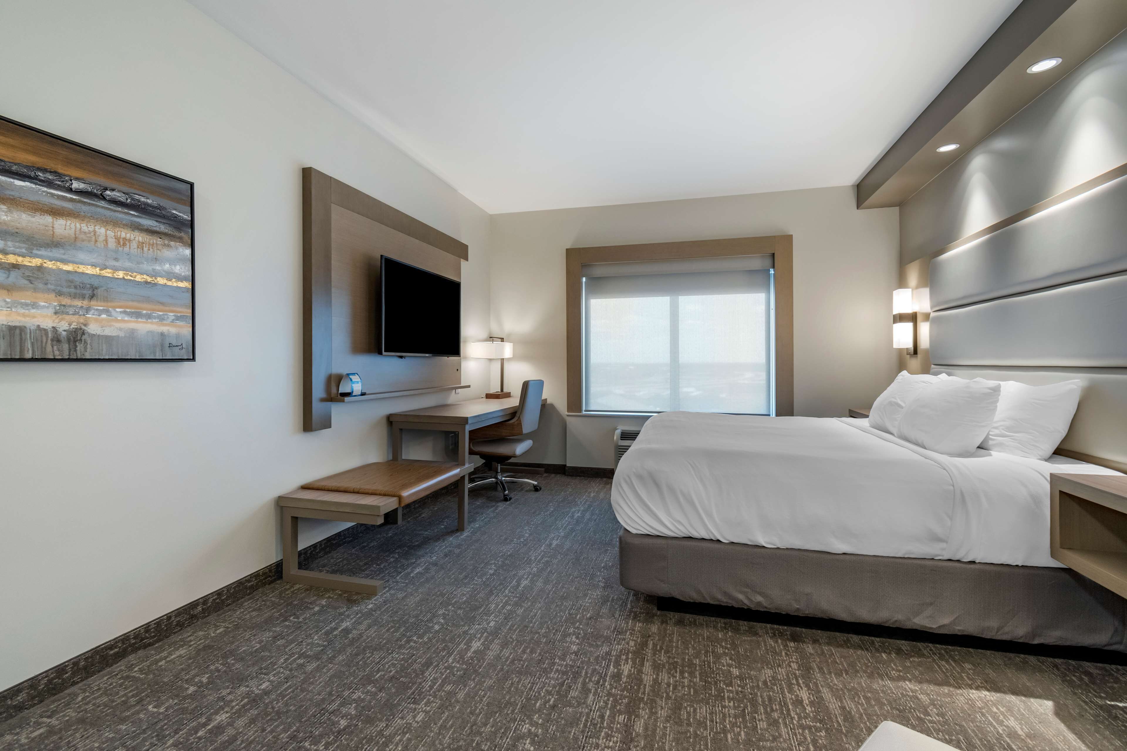 A hotel room with a bed, desk, chair, TV, and artwork on the wall.