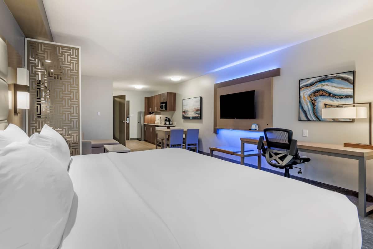 Ein Hotelzimmer mit einem großen Bett, einem Schreibtisch mit Stuhl, einer Küchenzeile und einem an der Wand befestigten Fernseher.