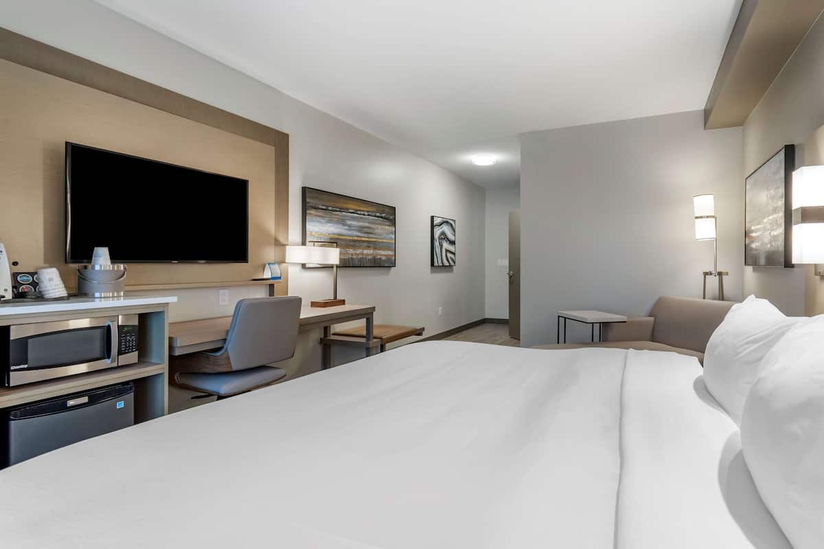 Ein Hotelzimmer mit einem großen Bett, einem Schreibtisch mit Mikrowelle, einem Sessel, einem Flachbildfernseher und einem kleinen Tisch mit Lampe.