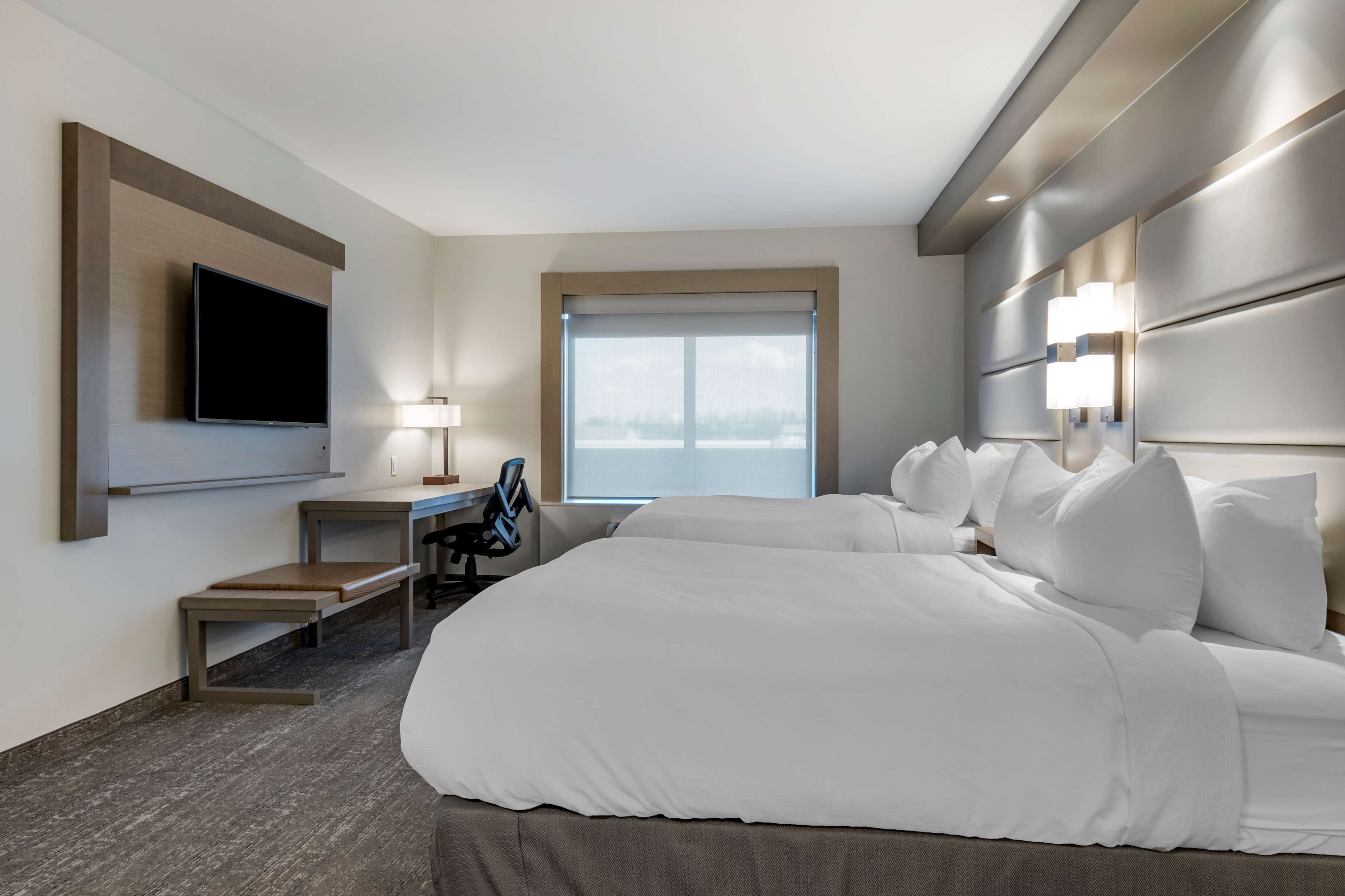 Ein Hotelzimmer mit einem großen Bett, einem Schreibtisch, einem Stuhl, einem Flachbildfernseher, einem Fenster mit Blick und einem mit Beleuchtung versehenen Kopfteil.