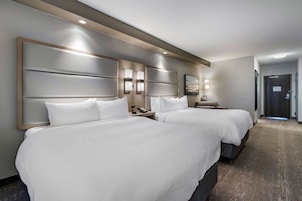 Habitación de hotel con dos camas, cabecero con iluminación integrada, mesita de noche con teléfono y puerta que da a un pasillo.