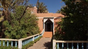 A pathway leading to a building with a traditional archway entrance, surrounded by lush greenery and palm trees. - Maison d'Hôtes au Jardin de Tamnougalt (Mezguita)
