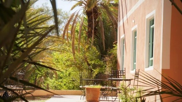 An outdoor patio with a table and chairs, surrounded by greenery and a building with a light-colored facade and green windows. - Maison d'Hôtes au Jardin de Tamnougalt (Mezguita)