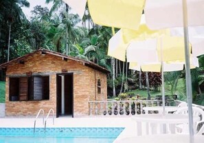 A small wooden cabin with a pool, surrounded by lush greenery and palm trees.