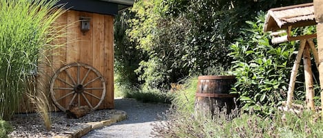 A rustic wooden building with a large wheel, surrounded by gravel paths and greenery.