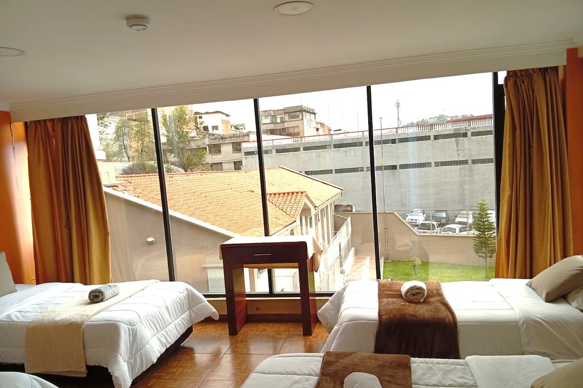 A hotel room with two beds, a small table, and a large window with a view of buildings and a bridge.