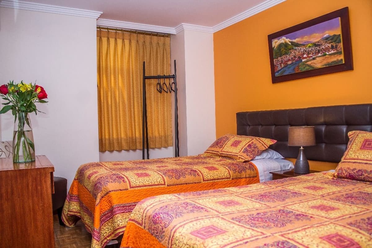 Une chambre d’hôtel avec deux lits, une fenêtre avec des rideaux, un tableau au mur et un vase de fleurs sur une table.